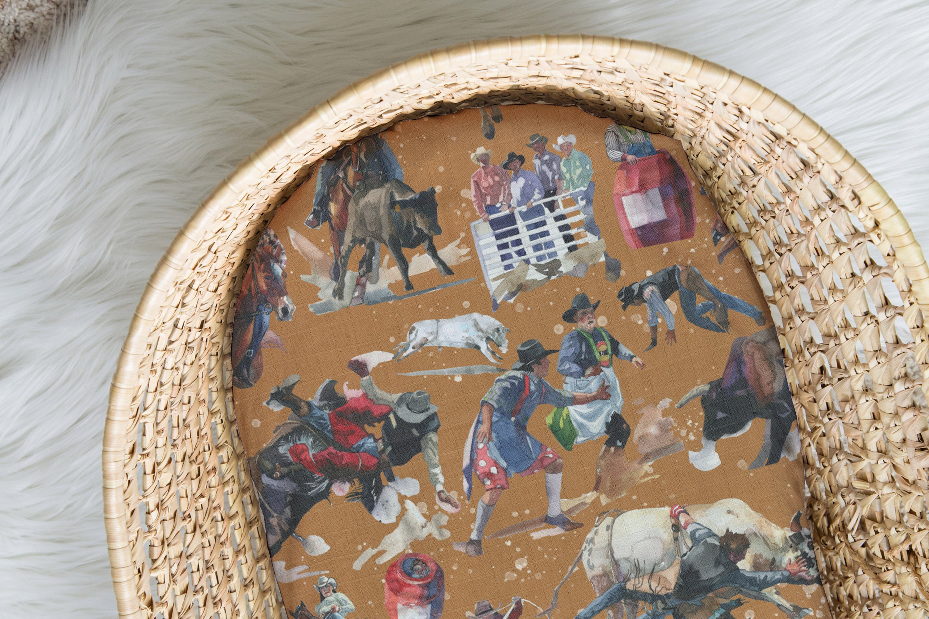 A tan-colored Moses basket sheet with a vintage rodeo-themed pattern, featuring images of cowboys, bulls, horses, and a rodeo clown.